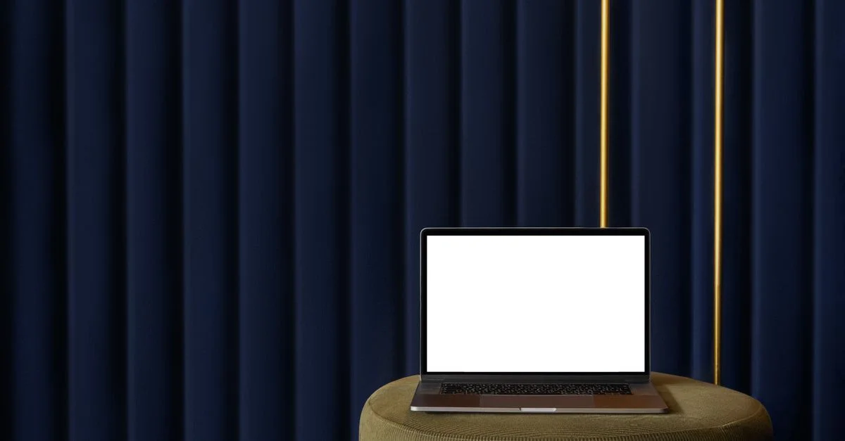 Un ordinateur portable moderne avec un écran vide est posé sur un tabouret vert contre un rideau bleu foncé.